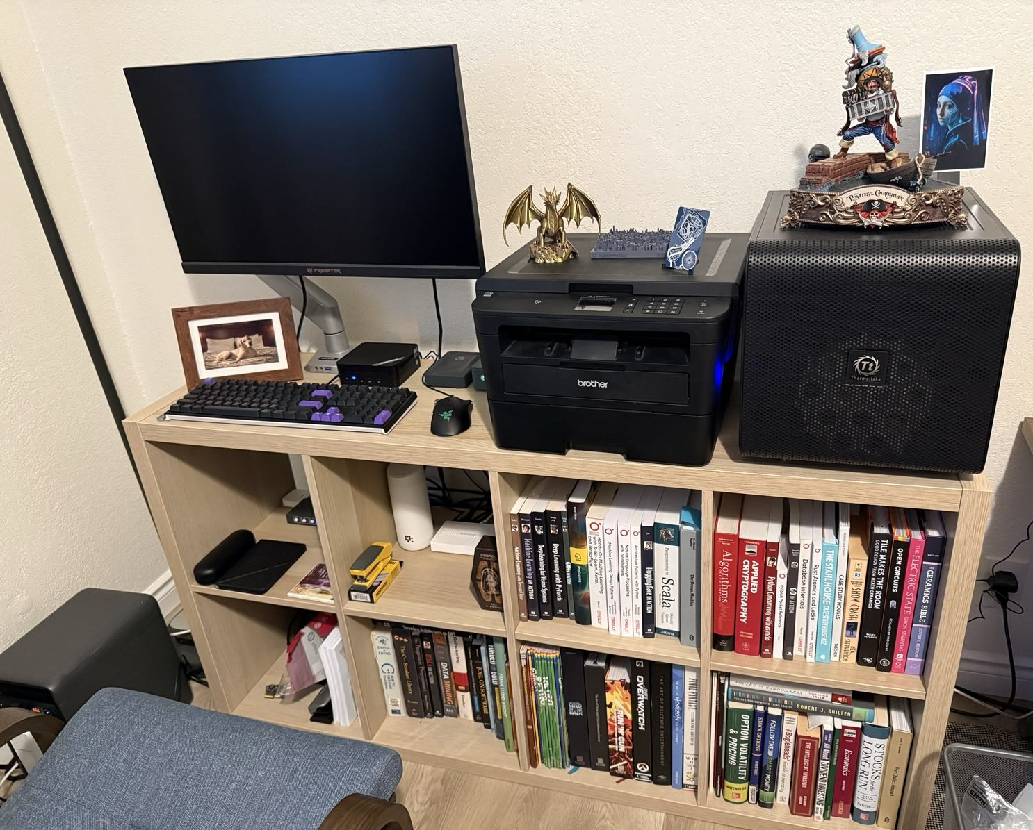 A shelf holding a monitor, mechanical keyboard, a mini-PC, a printer, and a Thermaltake tower case, above two rows of technical books.